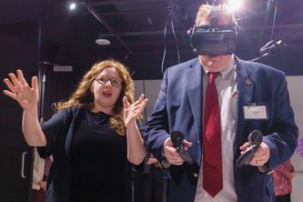 One person talking to another who is wearing a virtual reality headset and holding controls in each hand