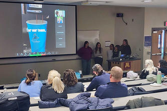 students watch a video presentation on a big screen at the front of a classroom
