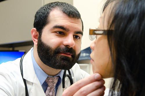 A bearded white male with a stethoscope examines examines a female Asian patient.