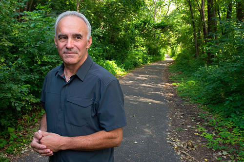 Cancer survivor Scott Nelson stands on a concrete path amongst a forest of trees with his hands crossed.