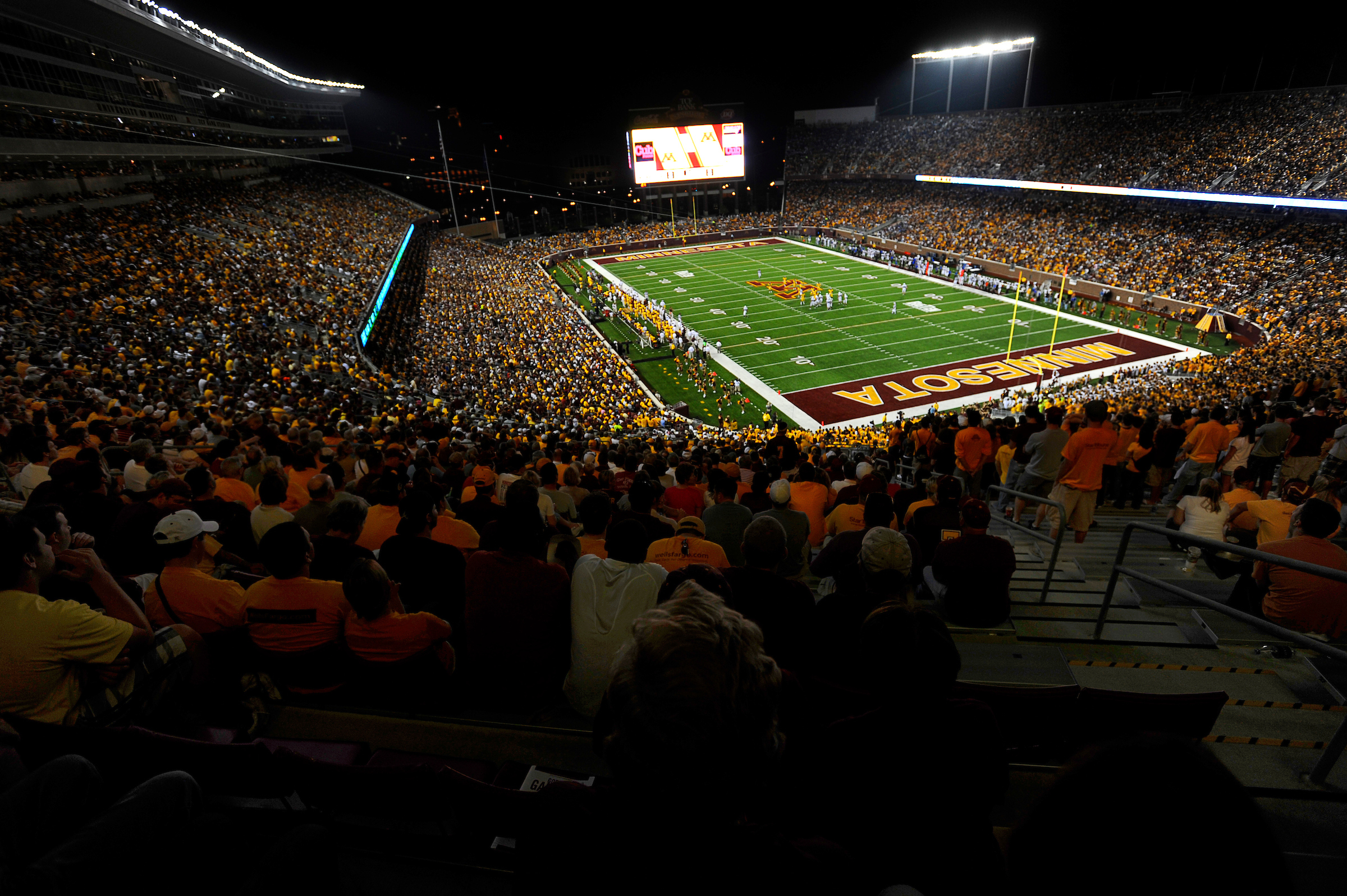 A wideangle shot of a capacity crowd watching a football game at night under the lights at Huntington Bank Stadium