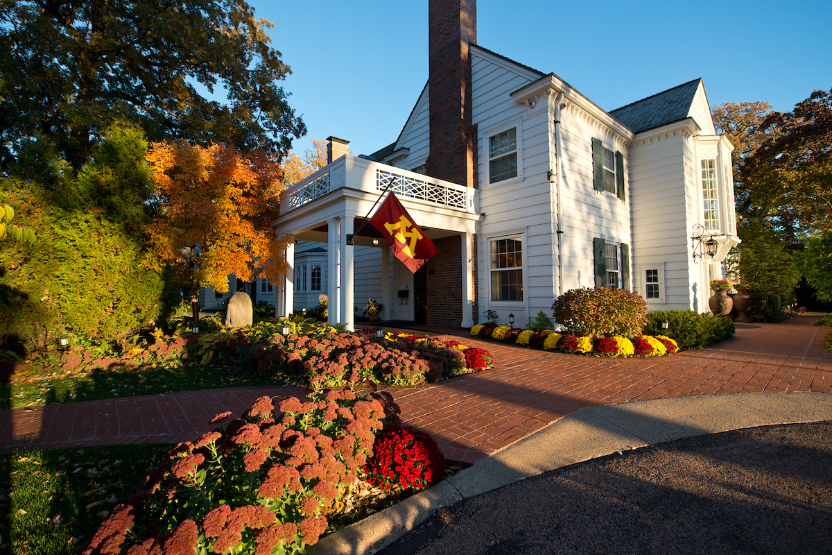 Evening sun casts long shadows over the front drive and white siding of the Eastcliff mansion, which is framed by fall foliage and a U of M flag waving near the entry.