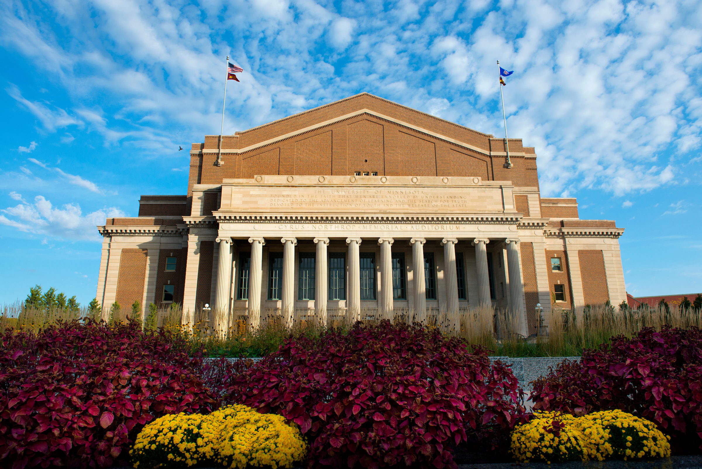 Northrop Auditorium against a blue sky with maroon and gold flowers in the foreground 
