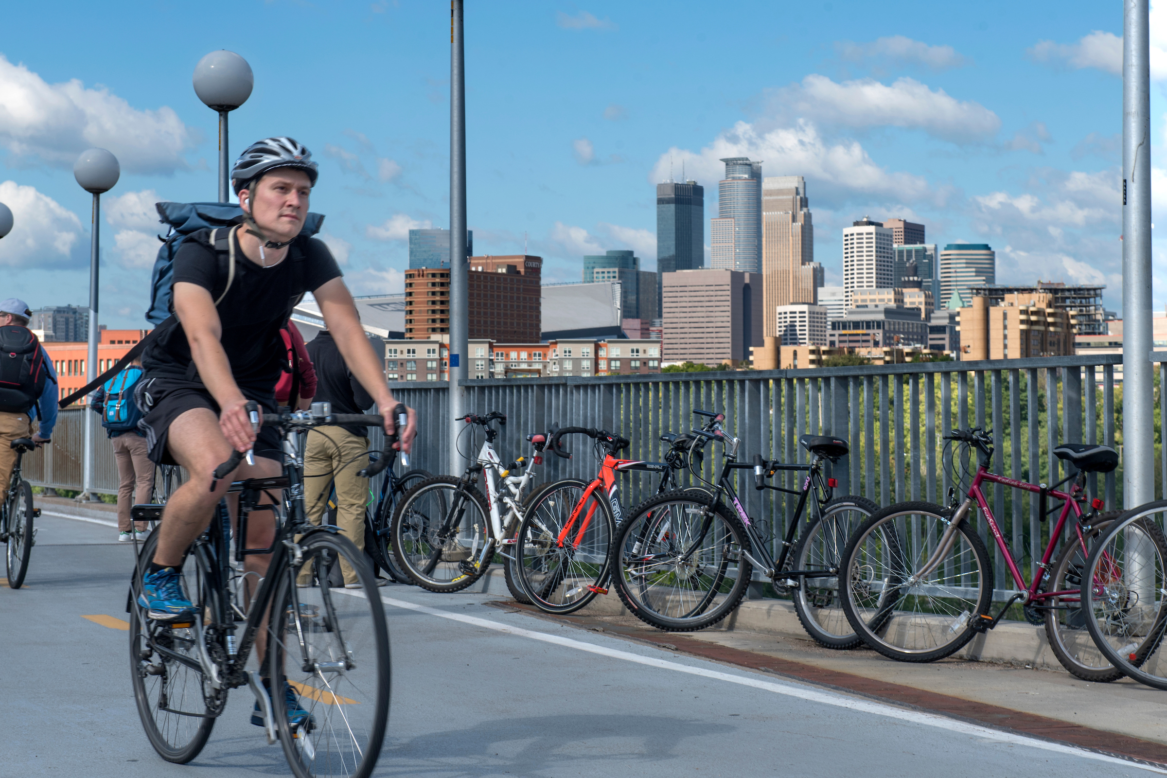 Man bikes across bridge, with a series of bikes parked against a fence in the background and the Minneapolis skyline rising above that.