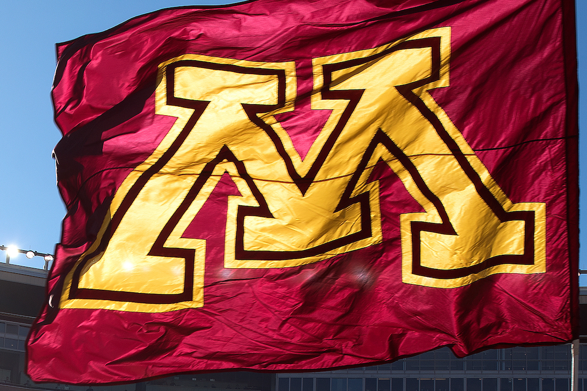 A windswept maroon flag with the U of M logo is backlit by sunshine.