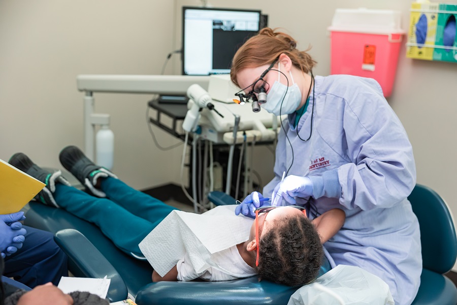 Image of a dentist in blue scrubs looking inside the mouth of a child who is laying on a chair receiving dental services.