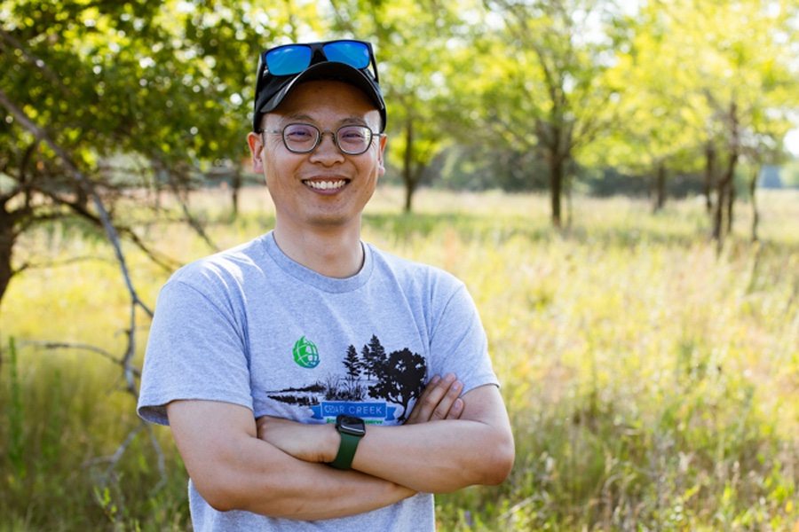 Maowei Liang, in glasses, T shirt and cap, with high grass and trees background.