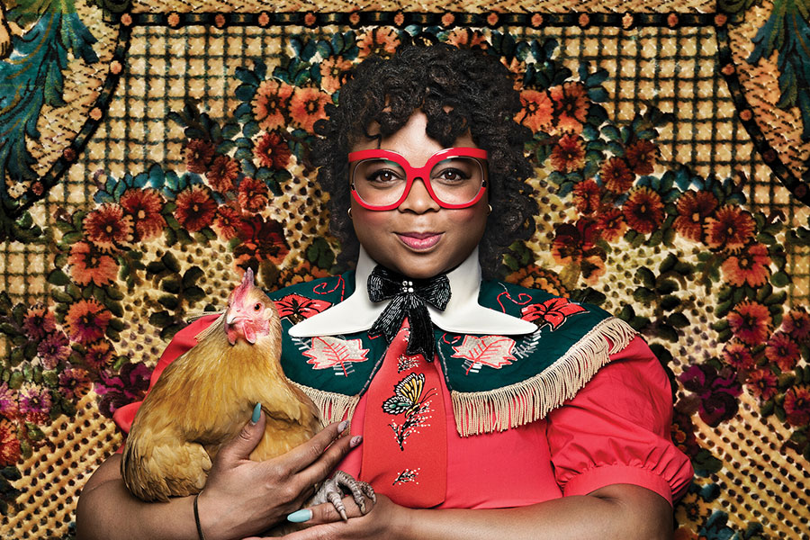 Myah Walker in a bright red dress holding a chicken with a decorative rug background. Photo by Shelly Mosman