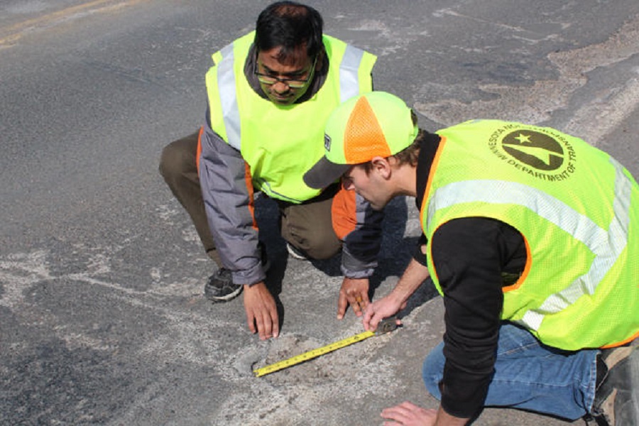 Image of Manik Barman and a student wearing a hat kneeling on the pavement using measuring tape to assess the size of a pothole while wearing reflective neon safety vests.