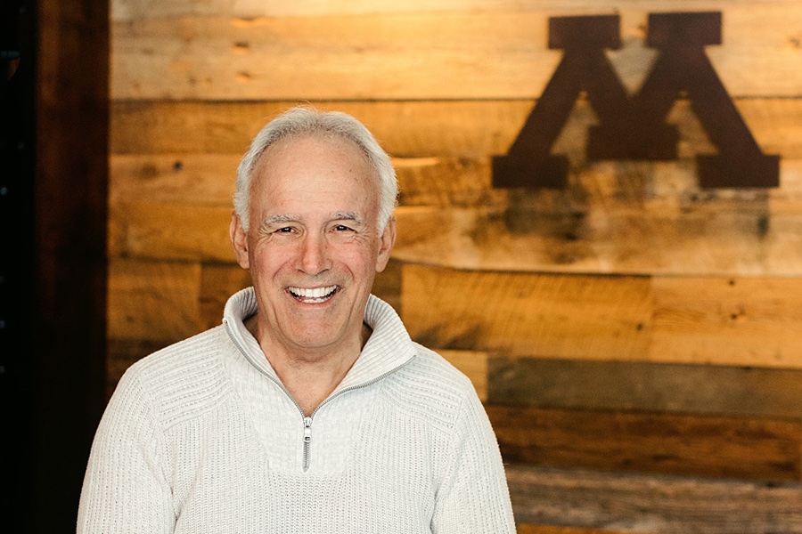 Scott Nelson, receding white hair, white hoodie and big smile, poses in front of a Big M against a wooden wall.