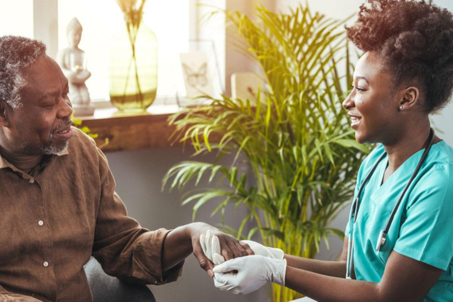 Black nurse wearing scrubs holding the hand of an elderly Black nursing home patient