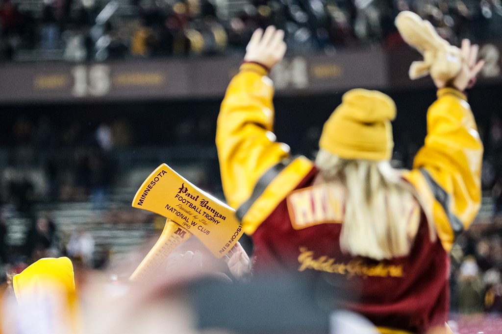 A Gopher fan on the shoulders of others raises her hands in a Maroon and Gold jacket, back facing the camera as she admires the Paul Bunyan Axe trophy