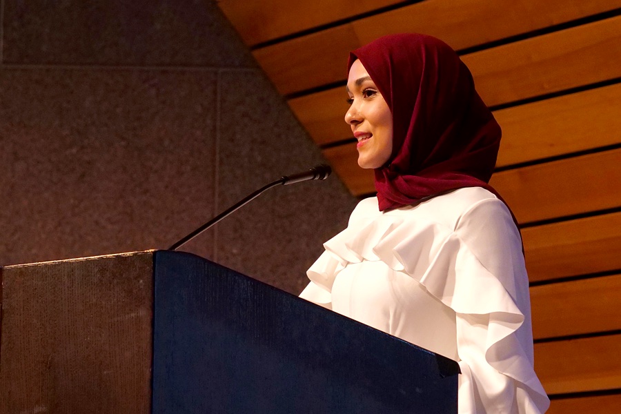 A young woman in maroon hijab and white dress, speaks at a lectern.