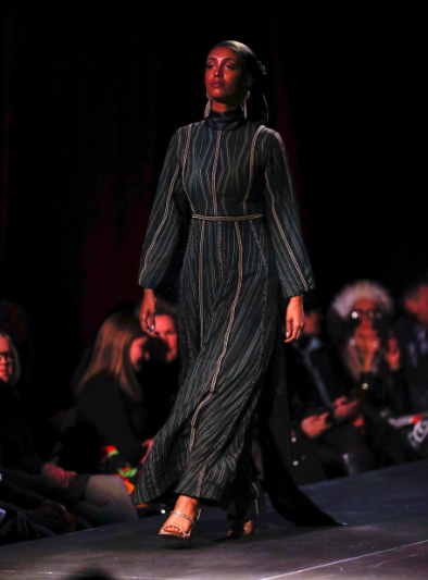 Model on runway during show wearing long-sleeved dark green long dress with vertical details