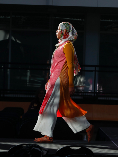 Somali models walks the runway in profile wearing white flowy pants, flower-print headscarf, and pink and orange flowy top