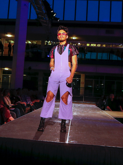Model posing at end of runway during show, wearing light purple overalls with knee cutouts over stiped half-shirt, with boots and mink thin sunglasses