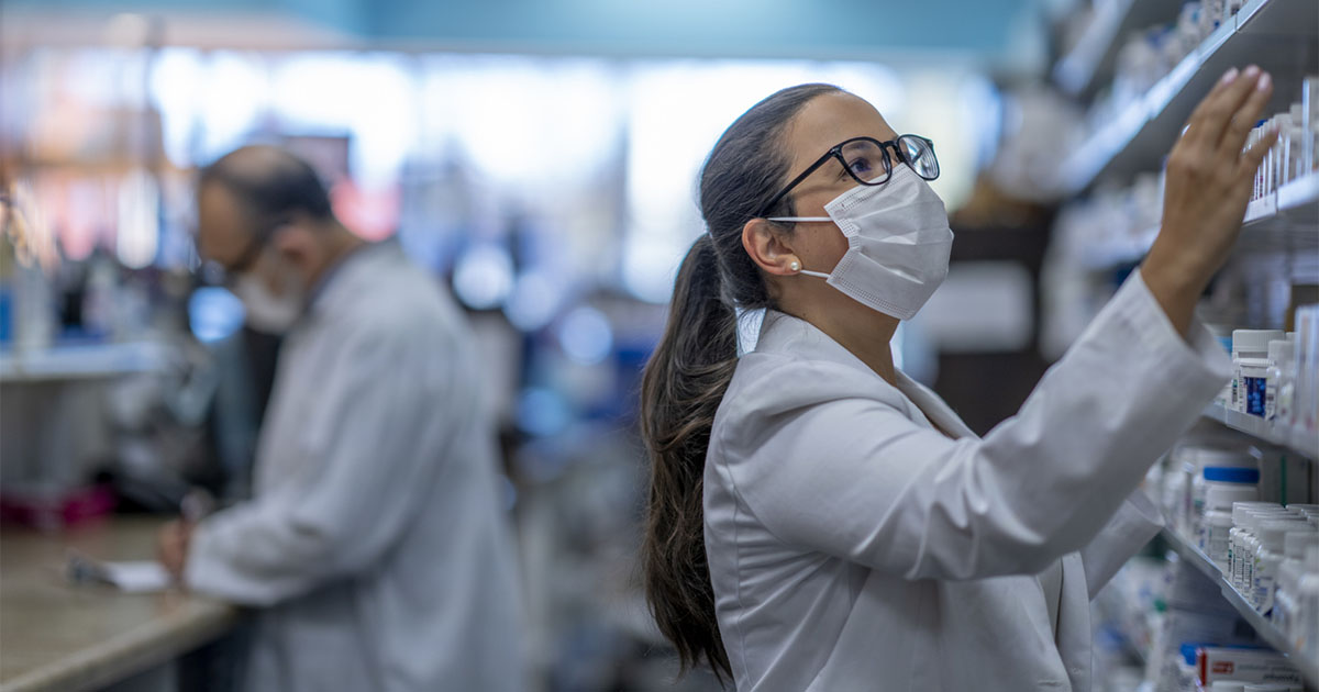 Woman wearing glasses, a face mask and a lab coat reaching for a bottle on a pharmacy shelf