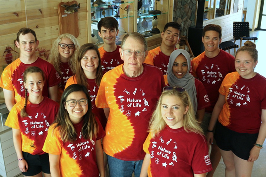 John. S. Anderson, a teacher with the first season of Nature of Life, poses with several students, all wearing identical orange and red shirts with what appears to be the stylized form of a Paramecium, a ubiquitous one-celled aquatic animal. 