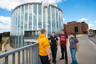 A University of Minnesota Police Department officer stands in from of the glass exterior of Bruiniks Hall, casually talking to three students