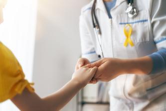 Image of doctor wearing yellow ribbon — which often represents sarcoma, or bone cancer — on their coat holding a child’s hand.