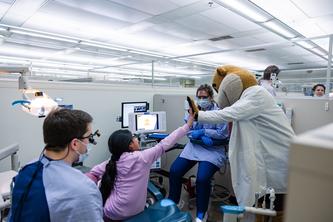 Image of the Goldy the Gopher mascot high fiving a child, surrounded by dentists in a clinic.