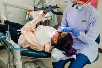 A girl wearing sunglasses reclines in a dental chair and holds a mirror as she has her teeth cleaned.
