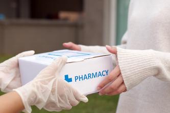 a close-up of hands wearing medical gloves handing a box labeled "Pharmacy" to a woman. 