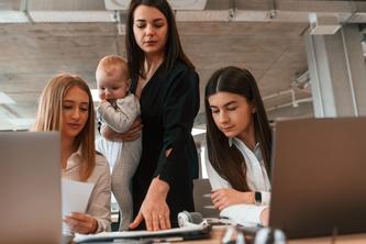 A woman stands with a baby between two seated women working at computers.