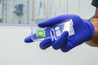 Image of a hand with a latex glove holding a computer chip in a plastic container.