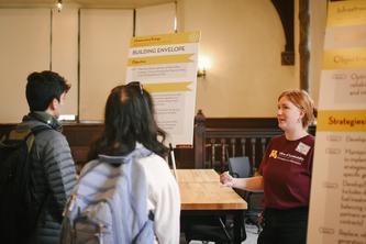 A woman with a maroon Office of Sustainability T-shirt on the right side speaking to two students on the left. Between the woman and students is a billboard titled "Building Envelope" on a table. 