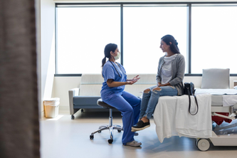Patient siting on a bed in a discussion with their doctor in scrubs sitting on a nearby stool