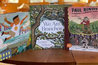 Three children’s books on a wooden table. The books are: “We are Branches,” “I'm From,” and “Paul Bunyan: The Invention of an American Legend.” 