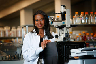 Abygail Andebrhan standing next to a microscope wearing a white coat