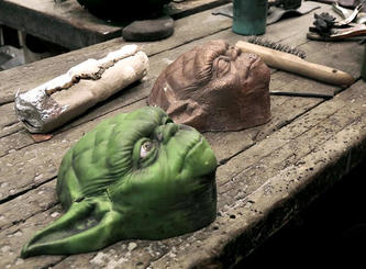 A green Yoda mask and a bronze cast of it sit on a workbench