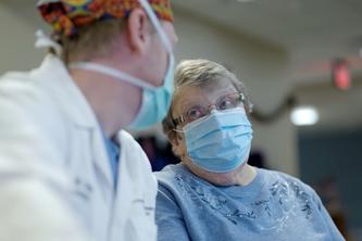 Mary Lou Peterson, gray hair and surgical mask, and Dr. Andy Grande, in white coat, mask and colorful cap, regard each other.