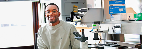A black male student smiling in a lab leaning on a desk with a microscope