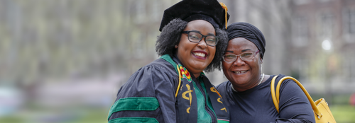 A black mother posing with her daughter wearing formal graduation clothing outside on campus