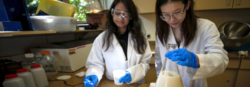 Two female students wearing gloves and lab coats mixing chemicals on a workbench with testing equipment
