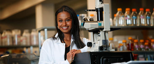 Abygail Andebrhan standing next to a microscope wearing a white coat