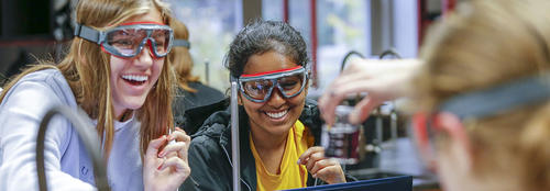 Two female students are laughing and smiling during a lab experiment wearing safety goggles