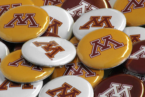 A collection of Maroon, Gold and white button-style pins adorned with the Block M representing the University of Minnesota are strewn in a pile