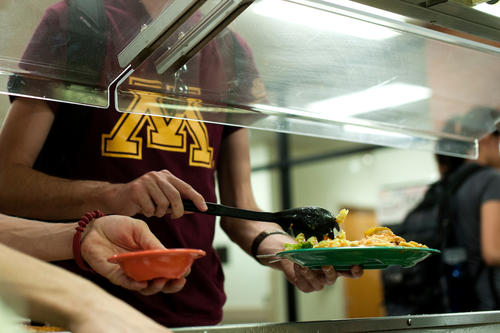 A student wearing a Maroon shirt with a Gold Block M dishes up a plate of food under the sneeze guard at a University dining facility
