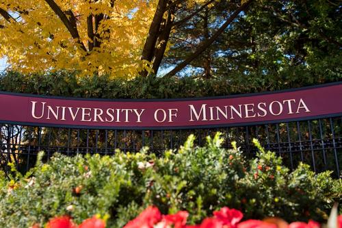 A black, wrought-iron gate, set back into landscaping, showcases a Maroon and Gold "University of Minnesota" sign that welcomes visitors to the Twin Cities campus