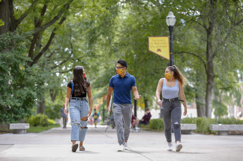 Three students, wearing masks to mitigate the spread of COVID-19, talk as they walk together along the sidewalks of the Northrop Mall on the Twin Cities campus
