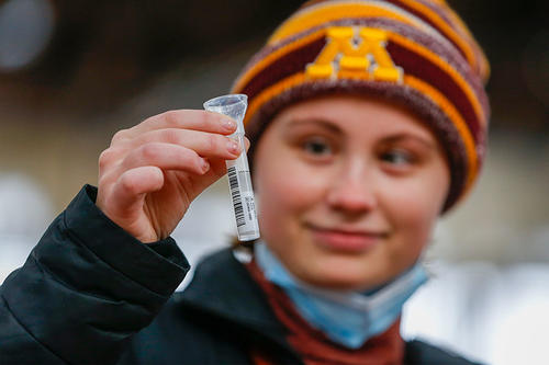 A young woman with an M stocking cap holds up her saliva sample.