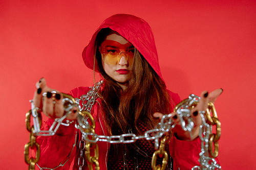 Carmen Aguirre, in red hooded cloak and long brown hair, holds a chain in outstretched hands. 
