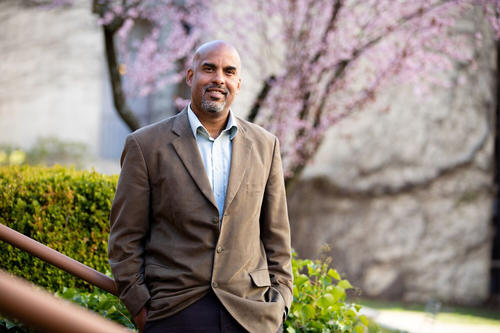 Damien Fair, bald, bearded and Black, wears a brown suit jacket and stands before a flowering tree.
