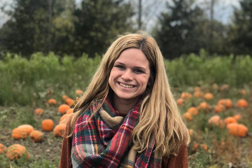 Hannah Pichman, long blonde hair and plaid scarf, stands in a pumpkin patch.