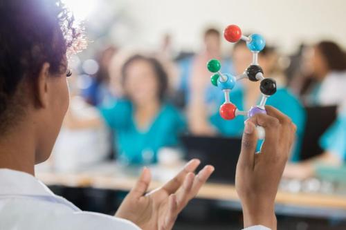 Stock photo of a Black female professor showing a class a model of molecular structure. The students are blurred in the background.