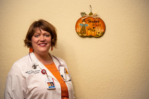 U of M Medical School Assistant Professor Beth Thielen poses for a photo in an orange shirt and white lab coat, in front of a beige wall with a pumpkin-shaped wall decoration that reads "Happy Fall"